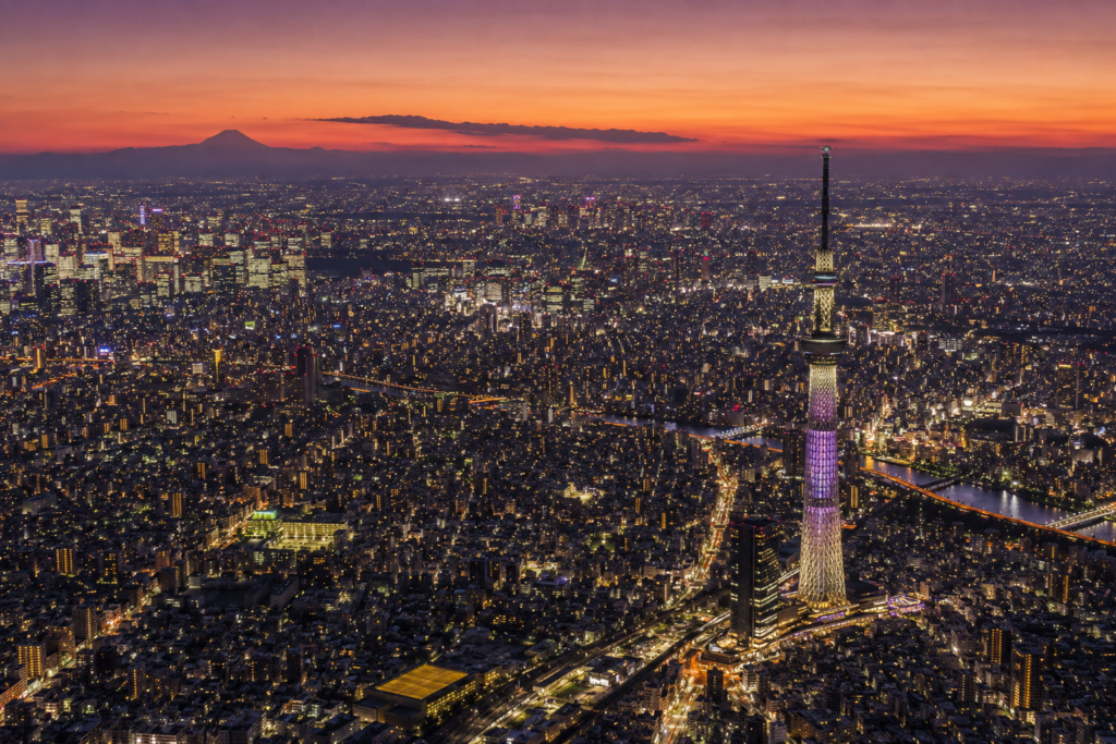 Tokyo travel guide feature image — aerial dusk view of Tokyo showing the illuminated Tokyo Skytree tower above the vast urban grid, the Sumida River threading through the city, the neon glow of Shinjuku on the horizon and Mount Fuji silhouetted in dusky purple representing the extraordinary scale of the world's most visited city