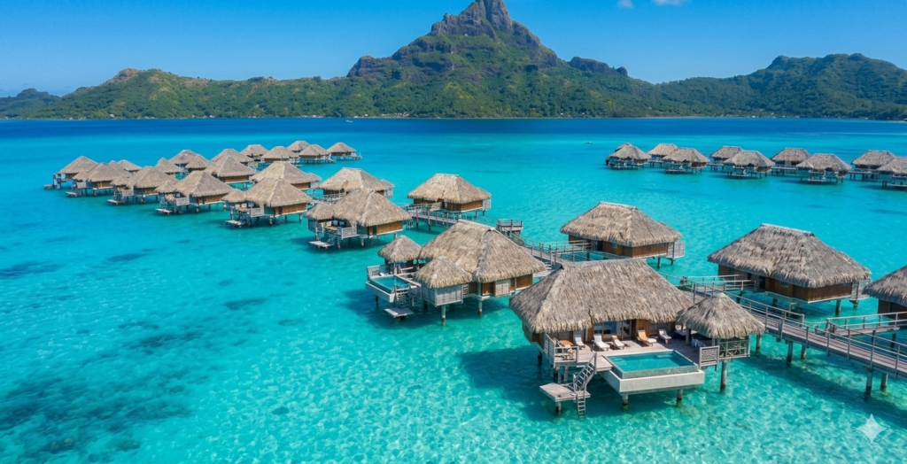 Aerial view of luxury overwater bungalows stretching over a crystal-clear turquoise lagoon in Bora Bora, French Polynesia, with a volcanic mountain in the background under a blue sky.