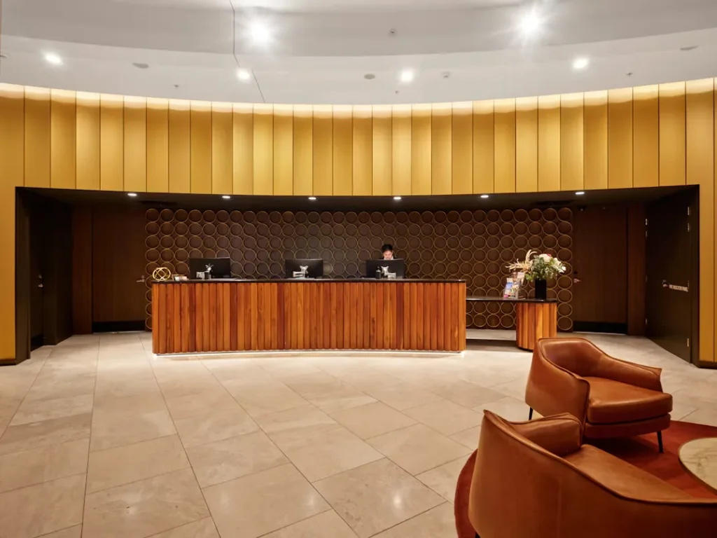 Staff member behind a wooden hotel reception desk with chairs in the lobby